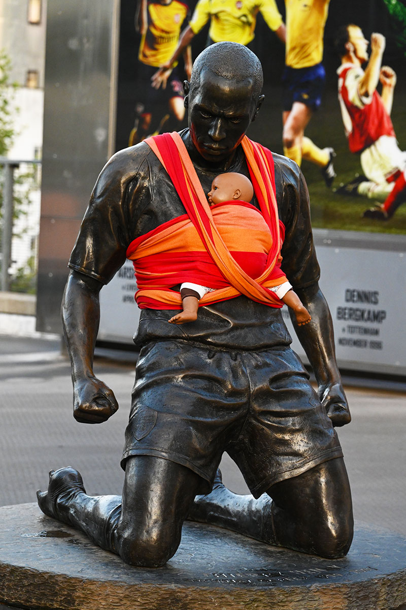 Statue of former Arsenal player Thierry Henry in front of Emirates Stadium in London with a red baby sling and baby doll on his chest